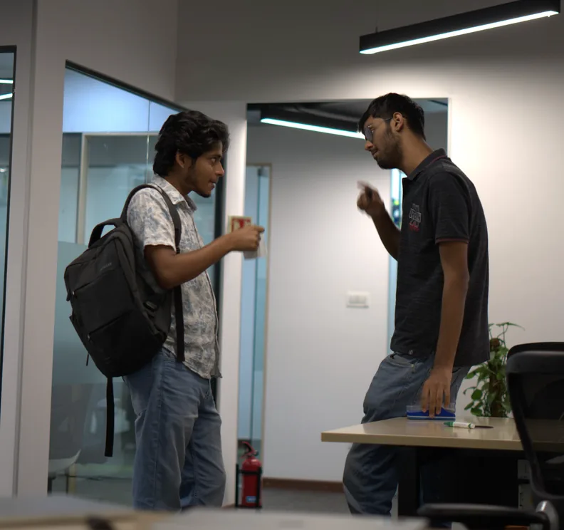 Two team members in conversation in a corridor, one with a backpack and a cup in hand.