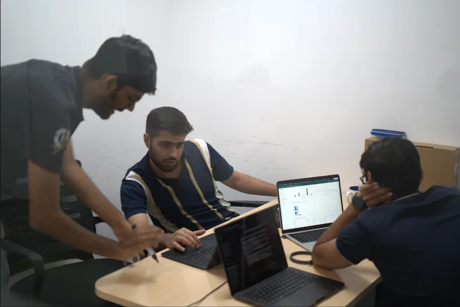 Three team members around a desk with laptops, one leaning in to point at a screen.